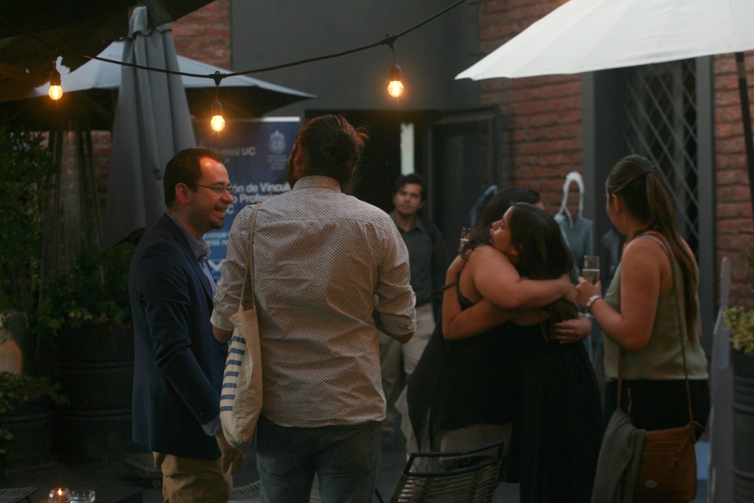 Personas conversando y compartiendo en un evento social al aire libre por la tarde, con luces colgantes, copas en mano y un abrazo entre dos asistentes. Al fondo, un cartel con el logo de Alumni UC.