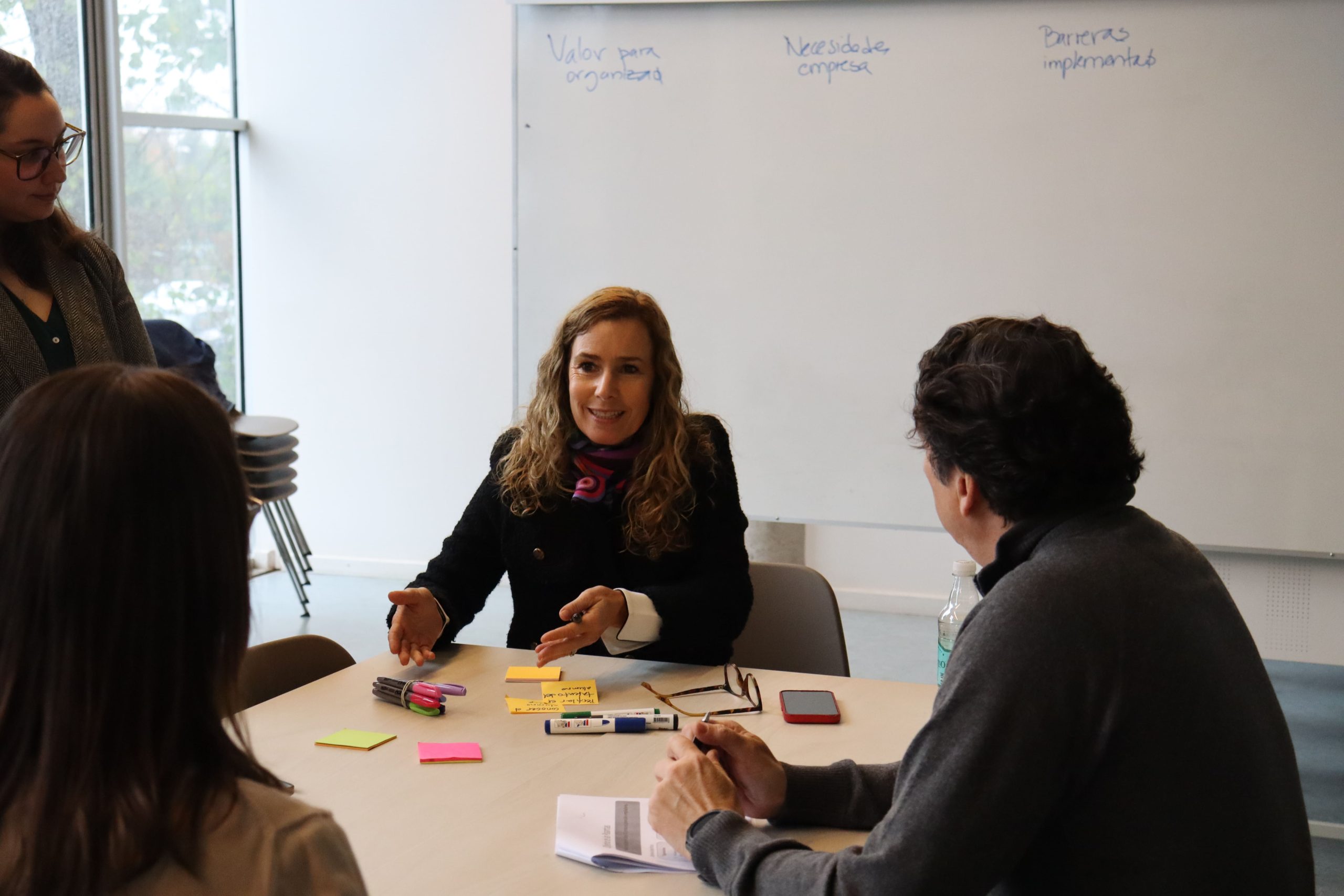 Cuatro personas, tres sentadas y una de pie, dialogan en torno a una mesa en una sala con luz natural. La mujer en el centro sonríe mientras habla y usa las manos para expresarse. En la mesa hay post-its de colores, marcadores y documentos. Detrás de ellos hay una pizarra blanca con frases escritas, como “Valor para organización”, “Necesidad empresa” y “Barreras implementación”.