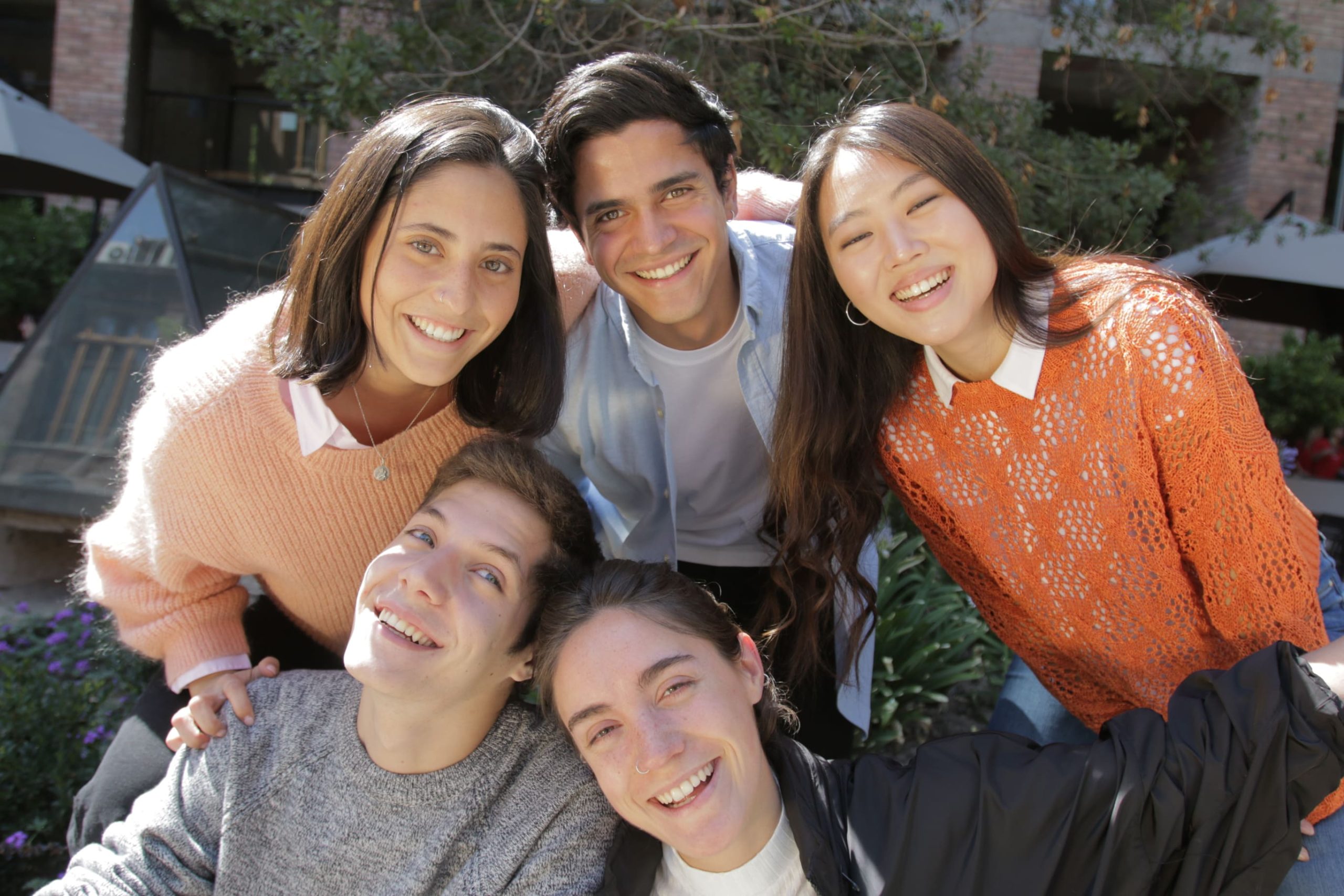 Cinco jóvenes sonríen al aire libre, en un entorno con vegetación y edificios de ladrillo al fondo. Están agrupados, inclinados hacia la cámara. Dos personas están al frente, sentadas o agachadas, y tres detrás, de pie. Visten ropa casual de colores claros y cálidos, proyectando un ambiente alegre y amistoso.