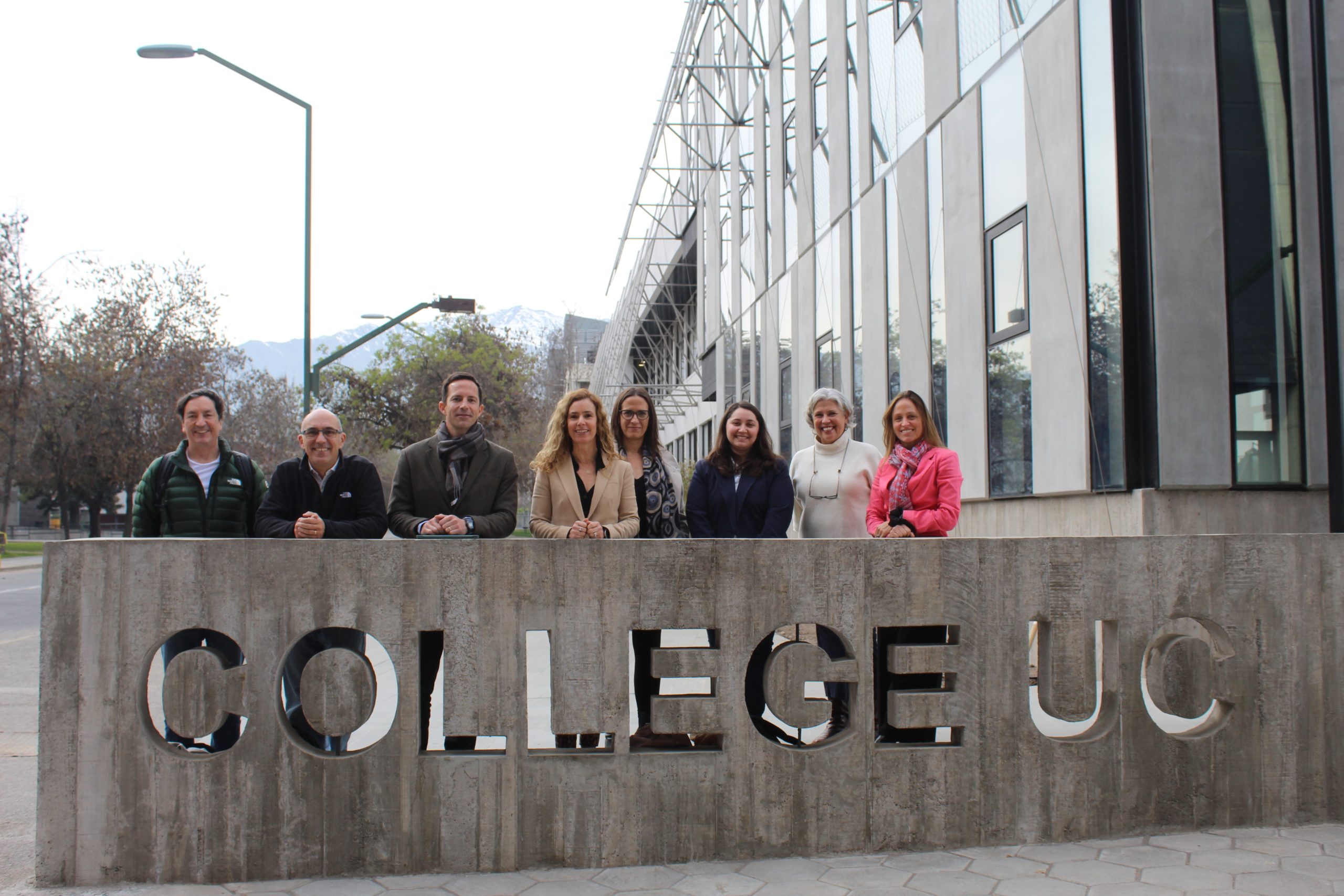 Equipo de College UC posando frente al letrero de hormigón con el nombre de la unidad académica en el Campus San Joaquín. Al fondo, se observa el edificio de College y la cordillera de los Andes.
