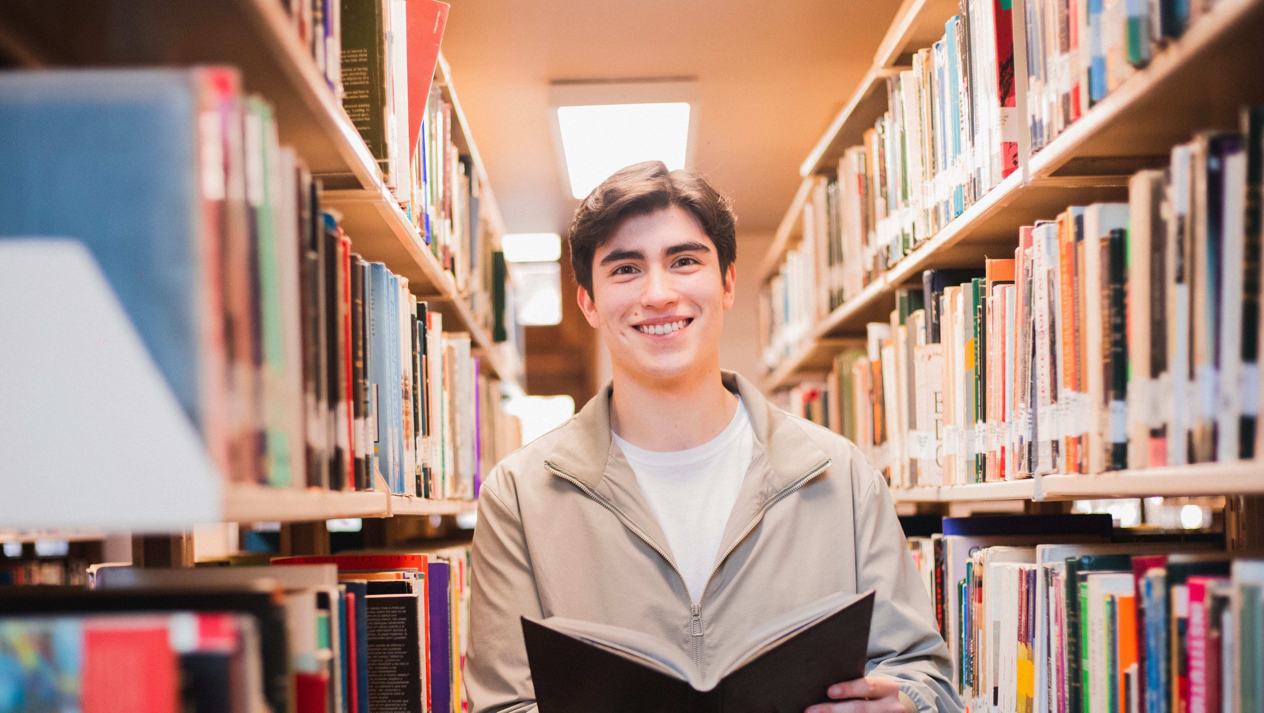 Estudiante de College UC sonríe en una biblioteca mientras sostiene un libro abierto entre estantes llenos de ejemplares.