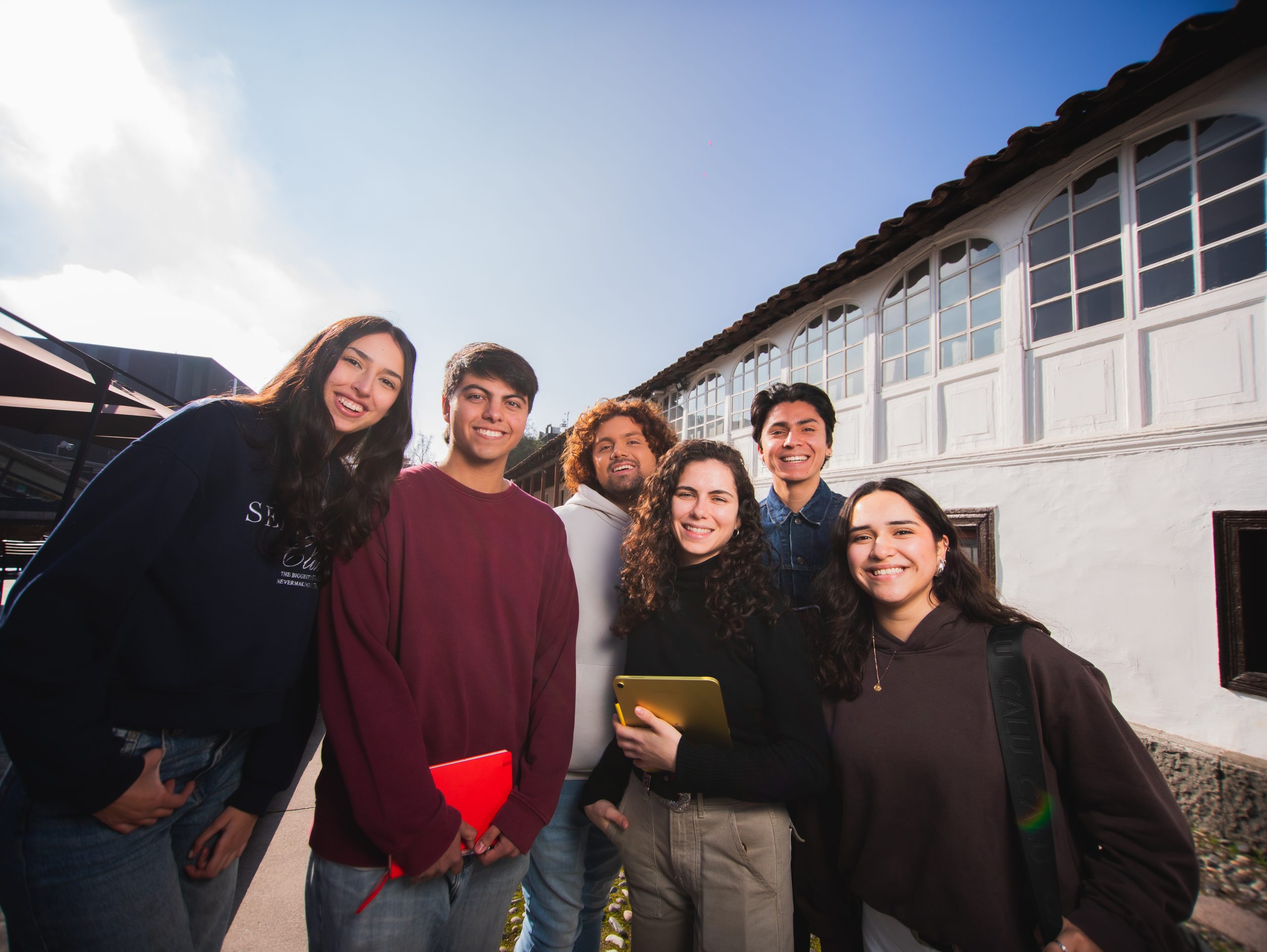 Grupo de seis estudiantes sonríe al aire libre frente a un edificio blanco con ventanales, en un día soleado